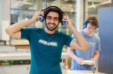 Decoratieve foto van een student die naar de camera lacht en zijn gehoorbescherming opdoet. Op de achtergrond is een medestudent aan het werken achter een werkbank. Ook hij draagt gehoorbescherming.