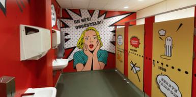 Illustratieve foto van de toiletten op het Houtens: de wanden en deuren zijn rood en geel en er zijn stripfiguren en kreten te zien, zoals 'Oh nee! Ongesteld!'